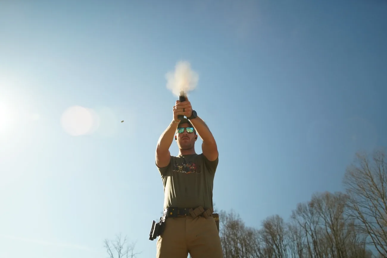A shooter firing a handgun at a low angle with sun flare behind the range